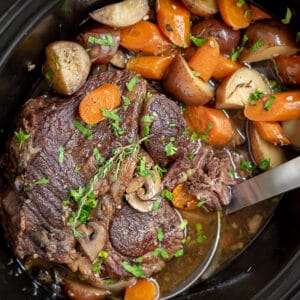 Crock pot filled with pot roast, potatoes, carrots, and mushrooms.