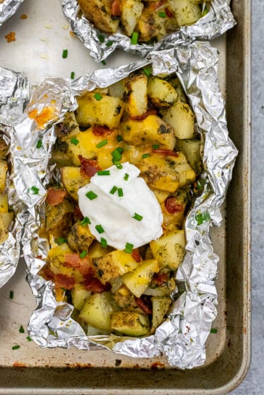 Sheet pan containing foil packets with cubed potatoes garnished with bacon, cheddar cheese, plain greek yogurt, and chives.