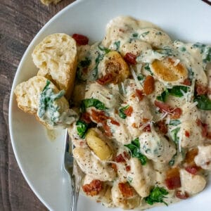 White bowl filled with creamy gnocchi, cooked baby spinach, and bacon. Served with a baked roll.