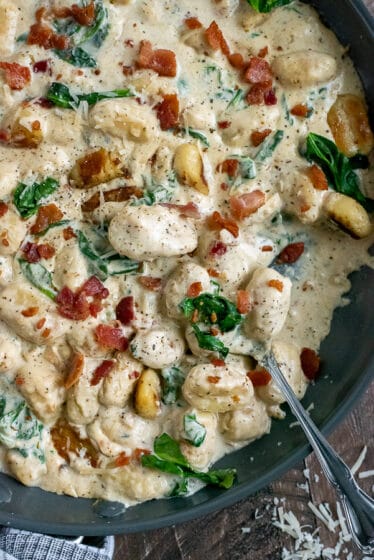 Black skillet filled with crispy gnocchi, cooked baby spinach, and bacon in a creamy parmesan sauce.