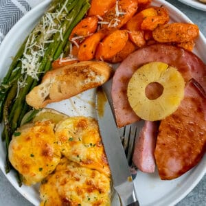 White plate filled with ham, cheesy potatoes, asaparagus, and carrots.